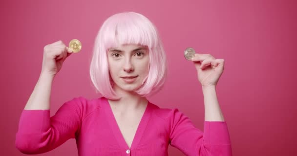 Junges weibliches Modell zeigt Bitcoin. Statische Aufnahme einer jungen Frau mit hellen Haaren, die Bitcoin vor der Kamera demonstriert, während sie vor rosa Hintergrund steht - Filmmaterial, Video