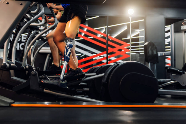 Sports woman with artificial leg running on treadmill at gym. Woman with prosthetic leg using walking on treadmill while working out in gym. - 写真・画像