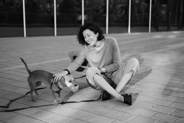 Jonge vrouwelijke volwassene in de stad met een hond zittend en knuffelend, lachend. Het aaien van de hond, voeden, plezier hebben Hoge kwaliteit foto - Foto, afbeelding