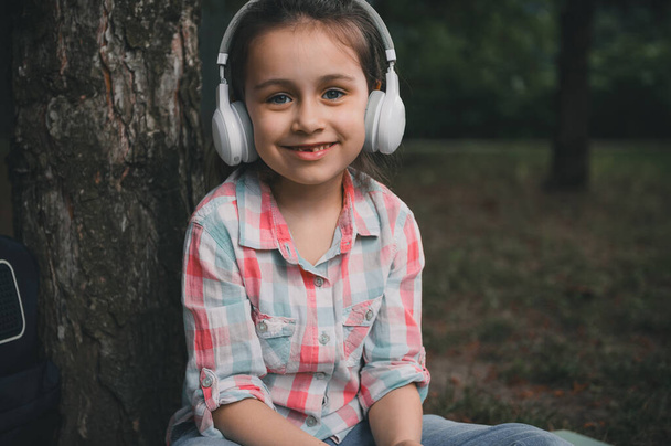 Крупним планом портрет кавказької щасливої маленької дівчинки, яка носить бездротові навушники, слухає музику, розслабляється після занять, сидить під деревом на траві і рівномірно посміхається дивиться на камеру - Фото, зображення