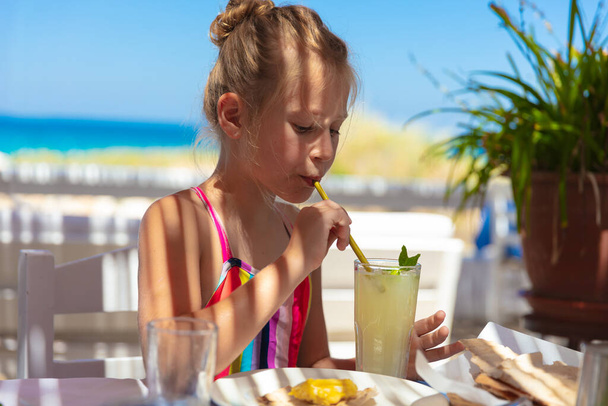Portrait of a cute little girl having a drink of a refreshing juice in the beach restaurant. Spending summer vacation on resort resort. - Fotó, kép