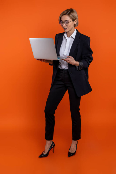 Confident woman entrepreneur wearing black official style suit working on notebook isolated over orange background. - Photo, image