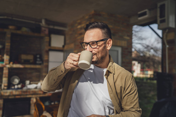 jeden dorosły biały mężczyzna stać na zewnątrz przed jego domem w słoneczny poranek lub dzień cieszyć się filiżanką herbaty lub kawy prawdziwi ludzie codziennie szczęśliwy uśmiech przestrzeń kopia - Zdjęcie, obraz