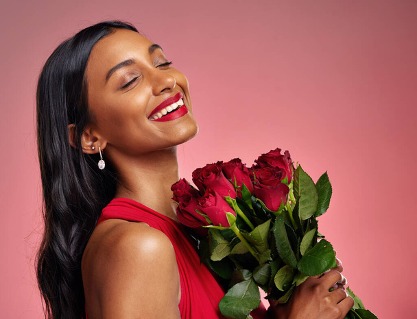 Πρόσωπο, ομορφιά και χαμόγελο μιας γυναίκας με τριαντάφυλλα σε φόντο στούντιο για την ημέρα του Αγίου Βαλεντίνου. Μακιγιάζ, μοντέλο και χαρούμενη νεαρή κοπέλα από την Ινδία με μπουκέτο λουλουδιών για ρομαντισμό ή αγάπη σε ροζ φόντο. - Φωτογραφία, εικόνα