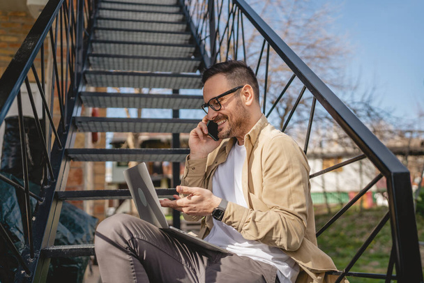 Один чоловік кавказький чоловік сидить на сходах на відкритому повітрі, приймаючи гальма від роботи з ноутбуком, використовуючи мобільний телефон, щоб зателефонувати розмовляючи щасливою посмішкою впевнена справжня людина - Фото, зображення