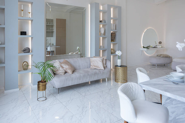 minimalistic light elegant luxury design of a modern spacious studio apartment with kitchen area, bedroom and huge mirror behind the couch. tiles on the floor and high ceiling. - Foto, imagen