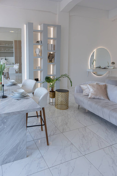 minimalistic light elegant luxury design of a modern spacious studio apartment with kitchen area, bedroom and huge mirror behind the couch. tiles on the floor and high ceiling. - 写真・画像