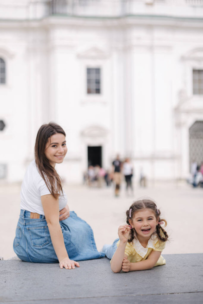 Portrait of pretty young woman with her adorable daughter sitting outdoor on stone bench. High quality photo - Foto, afbeelding