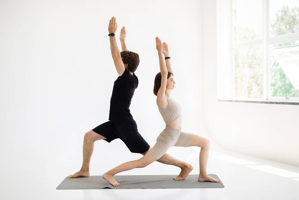Веселый молодой европейский парень и женщина в спортивной одежде делают упражнения для рук, практикуют йогу на коврике в гостиной, тренажерный зал интерьера. Спорт, активный образ жизни и уход за телом, совместные тренировки - Фото, изображение
