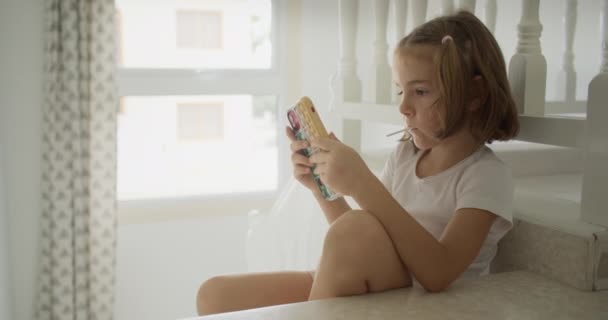 Het meisje is aan het bellen terwijl ze thuis op de trap zit. Een prachtig schattig kind kijkt video 's en sociale netwerken op een smartphone. Hoge kwaliteit 4k beeldmateriaal - Video