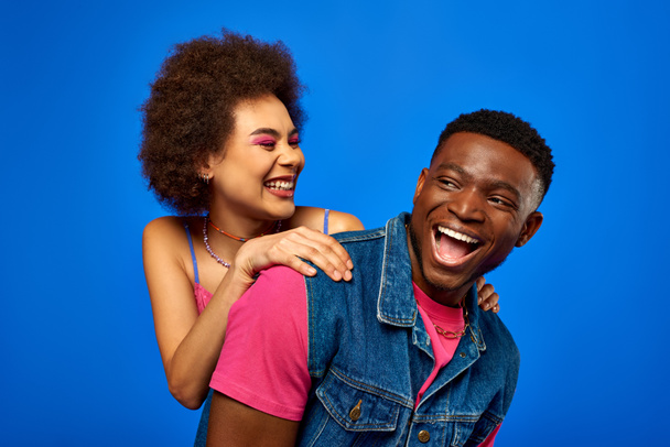 Smiling young african american woman with bold makeup hugging trendy best friend in summer outfit and spending time together isolated on blue, best friends in matching outfits, friendship - Photo, Image