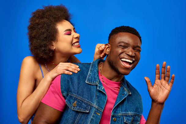 Θετική νεαρή Αφροαμερικανή γυναίκα με τολμηρό μακιγιάζ που αγγίζει το αυτί του στιλάτου καλύτερου φίλου με καλοκαιρινή στολή, ενώ διασκεδάζει και στέκεται απομονωμένη σε μπλε, καλύτερους φίλους με ασορτί ρούχα - Φωτογραφία, εικόνα