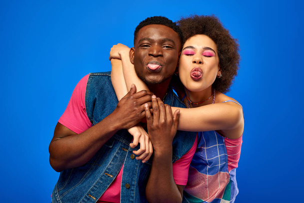 Позитивные молодые африканские лучшие друзья Америки в стильных ярких летних нарядах, торчащие языками и веселящиеся, стоя вместе изолированные на синих, лучшие друзья в одинаковых нарядах - Фото, изображение