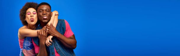 Jovens afro-americanos melhores amigos em roupas de verão brilhantes e na moda abraçando e salientando línguas enquanto estão juntos isolados em azul, banner, melhores amigos em roupas combinando - Foto, Imagem