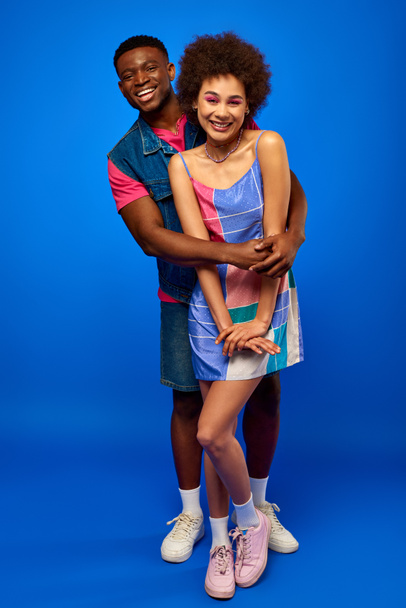 Full length of smiling african american man in bright summer outfit hugging stylish best friend in sundress while standing together on blue background, best friends in matching outfits - Foto, imagen