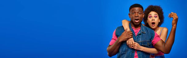 Geschokt jonge Afrikaanse Amerikaanse vrouw met gedurfde make-up knuffelen stijlvolle beste vriend in de zomer kleding en kijken naar camera geïsoleerd op blauwe, modieuze vrienden in trendy kleding, banner  - Foto, afbeelding