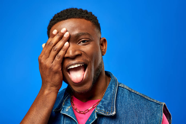 Excited young african american man in denim vest and bright t-shirt sticking out tongue and covering face while standing isolated on blue, trendy man showing summer style - Fotografie, Obrázek