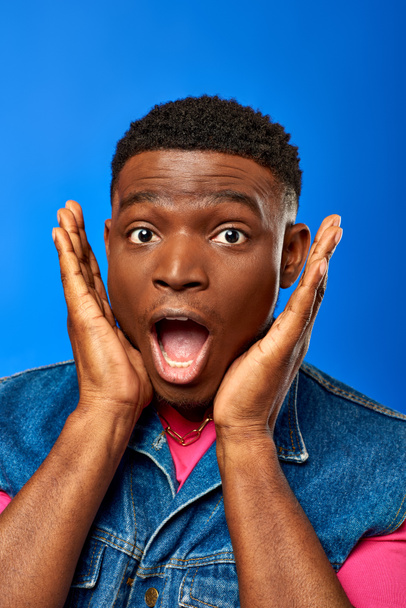 Portrait eines erstaunten jungen afrikanisch-amerikanischen Mannes mit moderner Frisur, der Jeansweste und rosa T-Shirt trägt, während er isoliert in die Kamera blickt und einen blauen, trendigen Mann zeigt, der Sommerstil zeigt - Foto, Bild
