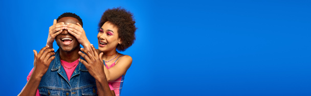 Sonriente mujer afroamericana joven con maquillaje brillante que cubre los ojos de la mejor amiga de moda en chaleco de mezclilla y de pie aislado en azul, amigos de moda en ropa de moda, pancarta  - Foto, imagen