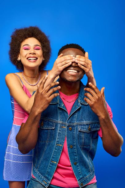 Positive young african american woman with bold makeup wearing sundress and covering eyes to best friend in summer outfit while standing isolated on blue, fashionable friends in trendy clothes - Foto, Bild