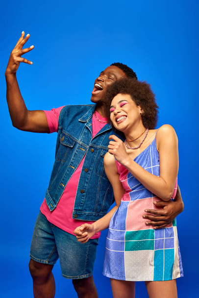Cheerful young african american man in denim vest and pink t-shirt hugging best friend in sundress and gesturing while standing isolated on blue, fashionable friends in trendy clothes - Photo, Image