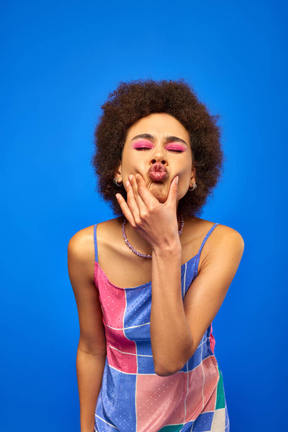 Portrait de jeune femme afro-américaine élégante avec maquillage audacieux et robe de soleil fermant les yeux et boudant les lèvres tout en posant et debout isolé sur bleu, modèle charismatique en tenue d'été - Photo, image