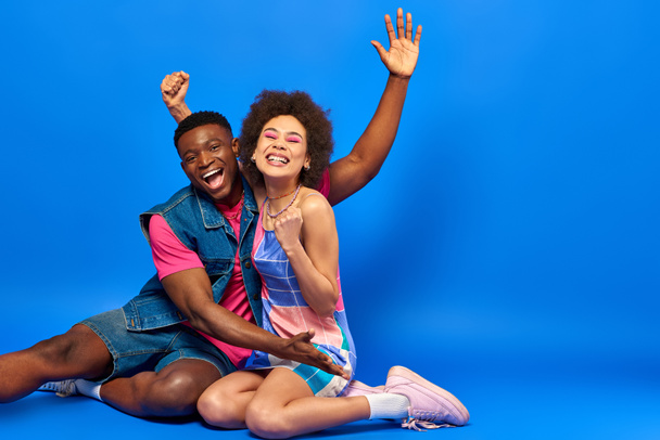 Cheerful young african american best friends in bright summer outfits waving hand and showing yes gesture at camera while sitting together on blue background, stylish friends posing confidently - Fotó, kép