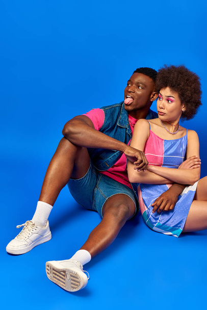Offended young african american best friends in bright summer outfits crossing arms and sticking out tongue while sitting together on blue background, stylish friends posing confidently - Photo, Image