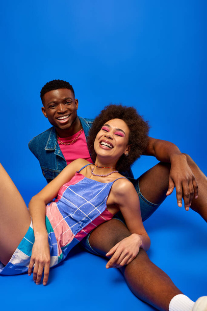 Neşeli ve modaya uygun Afro-Amerikan en iyi arkadaşları, parlak yaz kıyafetleri içinde, mavi arka planda birlikte poz verirken kameraya bakıyorlar.  - Fotoğraf, Görsel