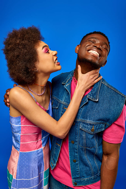 Enojada joven afroamericana mujer con maquillaje audaz tocando el cuello de alegre mejor amigo en traje de verano elegante, mientras que de pie aislado en azul, besties de moda irradiando confianza  - Foto, Imagen