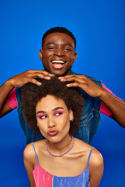 Glimlachende jonge Afro-Amerikaanse man met moderne kapsel en zomer outfit aanraken haar van beste vriend met gedurfde make-up geïsoleerd op blauwe, modieuze besties stralen vertrouwen, vriendschap - Foto, afbeelding