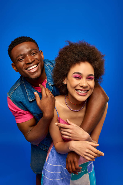 Cheerful young african american man in denim vest hugging best friend with bold makeup and stylish sundress while standing isolated on blue, fashionable besties radiating confidence  - Foto, Bild