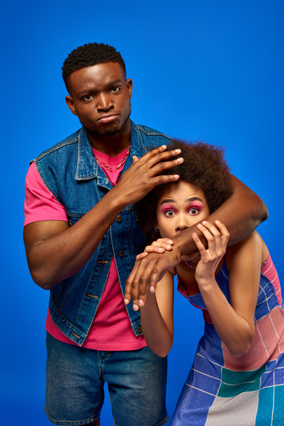 Serious young african american man in summer outfit covering scared best friend in stylish sundress and looking at camera isolated on blue, fashionable besties radiating confidence, friendship - Photo, Image