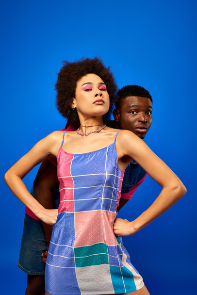 Confident young african american woman with bold makeup and sundress holding hands on hips while covering scared best friend isolated on blue, friends showcasing individual style, friendship - Valokuva, kuva
