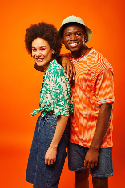Sonriente joven afroamericano con sombrero de panama y traje de verano mirando a la cámara cerca de mejor amigo con maquillaje audaz y de pie aislado en naranja, amigos mostrando el estilo individual - Foto, Imagen