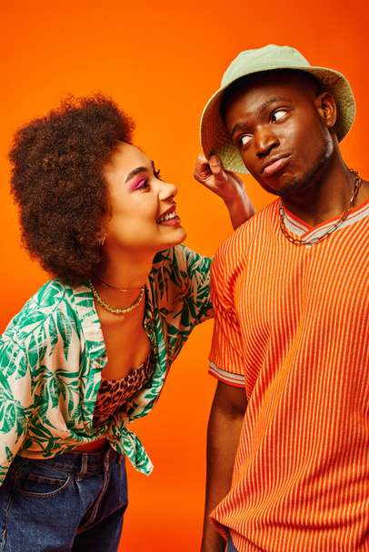 Χαρούμενη νεαρή Αφροαμερικάνα γυναίκα με τολμηρό μακιγιάζ αγγίζοντας panama καπέλο του μοντέρνου καλύτερου φίλου, ενώ περνούν το χρόνο τους απομονώνονται σε πορτοκαλί, φίλους προβάλλοντας ατομικό στυλ, φιλία - Φωτογραφία, εικόνα