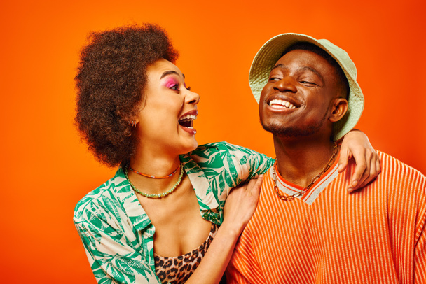 Возбужденная молодая африканская американка с смелым макияжем и модным костюмом, обнимающая лучшего друга в панамской шляпе и стоящая изолированная от оранжевого, друзья демонстрирующие индивидуальный стиль, дружбу - Фото, изображение