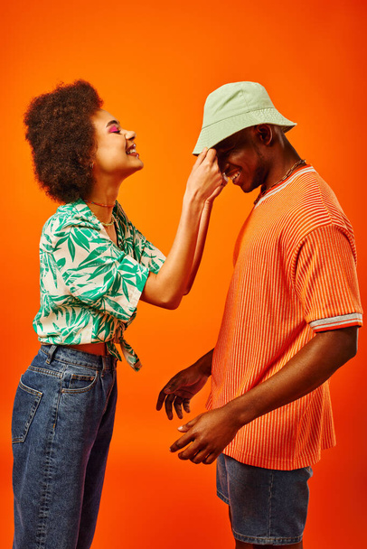 Yazlık kıyafetli, gülümseyen Afro-Amerikalı bir kadının en iyi arkadaşına şapkasını takması ve turuncu üzerinde tek başına dikilirken eğlenmesi. Moda kıyafetleri içinde arkadaşlar. - Fotoğraf, Görsel