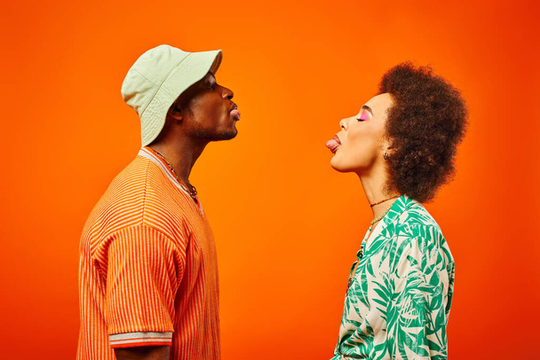 Side view of young and trendy african american best friends in summer outfits sticking out tongues while standing together isolated on orange, friends in trendy clothes, friendship concept - Foto, immagini