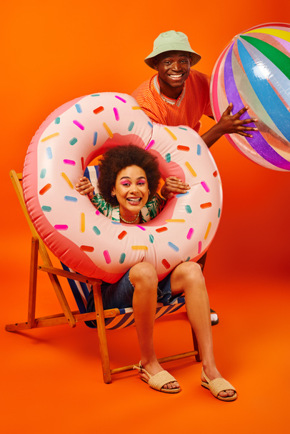 Pozitív fiatal afro-amerikai legjobb barátok nyári ruhák gazdaság medence gyűrű és labda, miközben ül a fedélzeten szék és a kamera narancssárga háttér, divat előre barátok - Fotó, kép