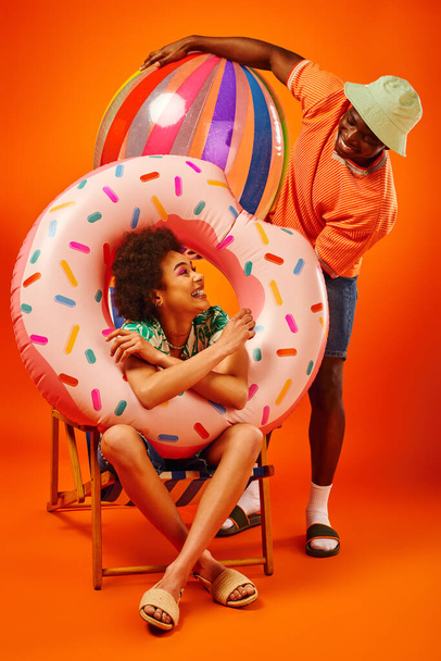 Vidám fiatal afro-amerikai nő nyári ruhában tartja medence gyűrű, miközben ül a fedélzeten szék és nézi a legjobb barát labda narancs háttér, divat előre barátok - Fotó, kép