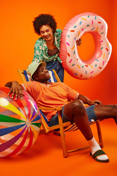 Mosolygó fiatal afro-amerikai nő nyári ruha gazdaság úszás gyűrű, miközben áll közel elegáns barátja panama kalap ül a fedélzeten szék narancssárga háttér, divat előre barátok - Fotó, kép