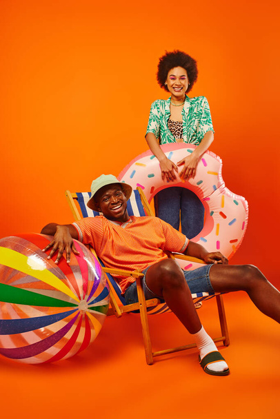 Pozitív fiatal afro-amerikai barátok nyári ruhák gazdaság úszás gyűrű és labda közelében nyugágy, miközben időt töltenek együtt narancs háttér, divat-előre barátok, barátság - Fotó, kép