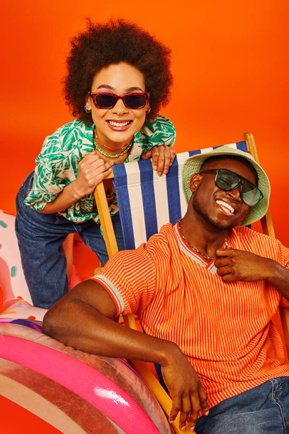 Pozitív fiatal afro-amerikai nő napszemüvegben és nyári ruha áll közelében legjobb barátja panama kalap a fedélzeten szék és strandlabda narancssárga háttér, divat előre barátok - Fotó, kép