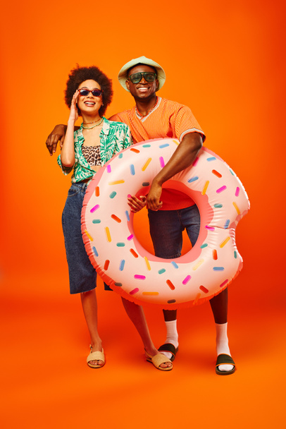 Volledige lengte van glimlachende jonge Afro-Amerikaanse man in zonnebril en panama hoed knuffelen stijlvolle beste vriend en het houden van zwemring op oranje achtergrond, vrienden in trendy casual kleding - Foto, afbeelding