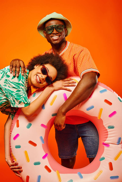 Positive young african american man in panama hat and sunglasses hugging trendy best friend in summer outfit and holding swim ring isolated on orange, friends in trendy casual attire - Foto, Bild