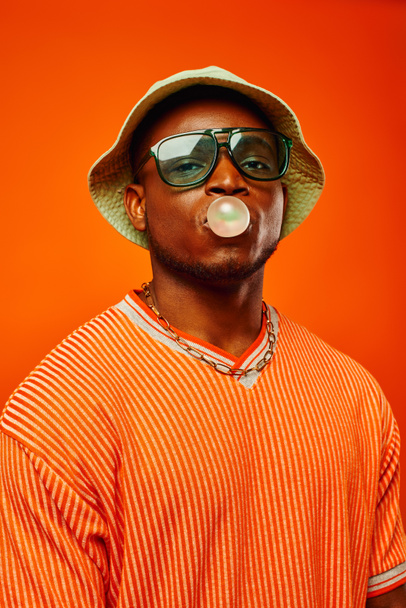 Portrait of trendy young african american man in sunglasses and panama hat blowing bubble gum and looking at camera while standing isolated on red, man with sense of style, confidence - Foto, imagen