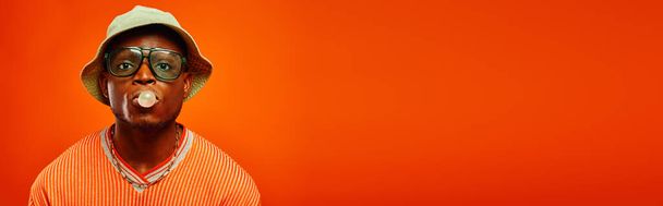 Giovane afroamericano alla moda in abito estivo, occhiali da sole e cappello panama che soffia gomma da masticare e guardando la fotocamera isolata sul rosso, uomo con senso dello stile, banner, fiducia - Foto, immagini