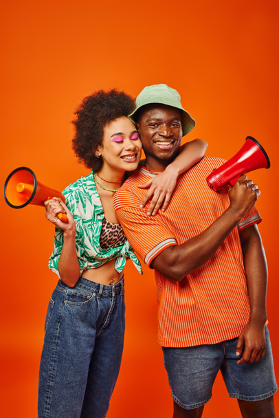 Smiling young african american woman with bold makeup and summer outfit hugging best friend in panama hat and holding loudspeaker while standing isolated on red, friends in fashionable outfits - Photo, Image