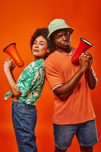 Confident young african american best friends in trendy summer outfits holding loudspeakers and looking at camera while standing isolated on red, friends in fashionable outfits - Photo, Image